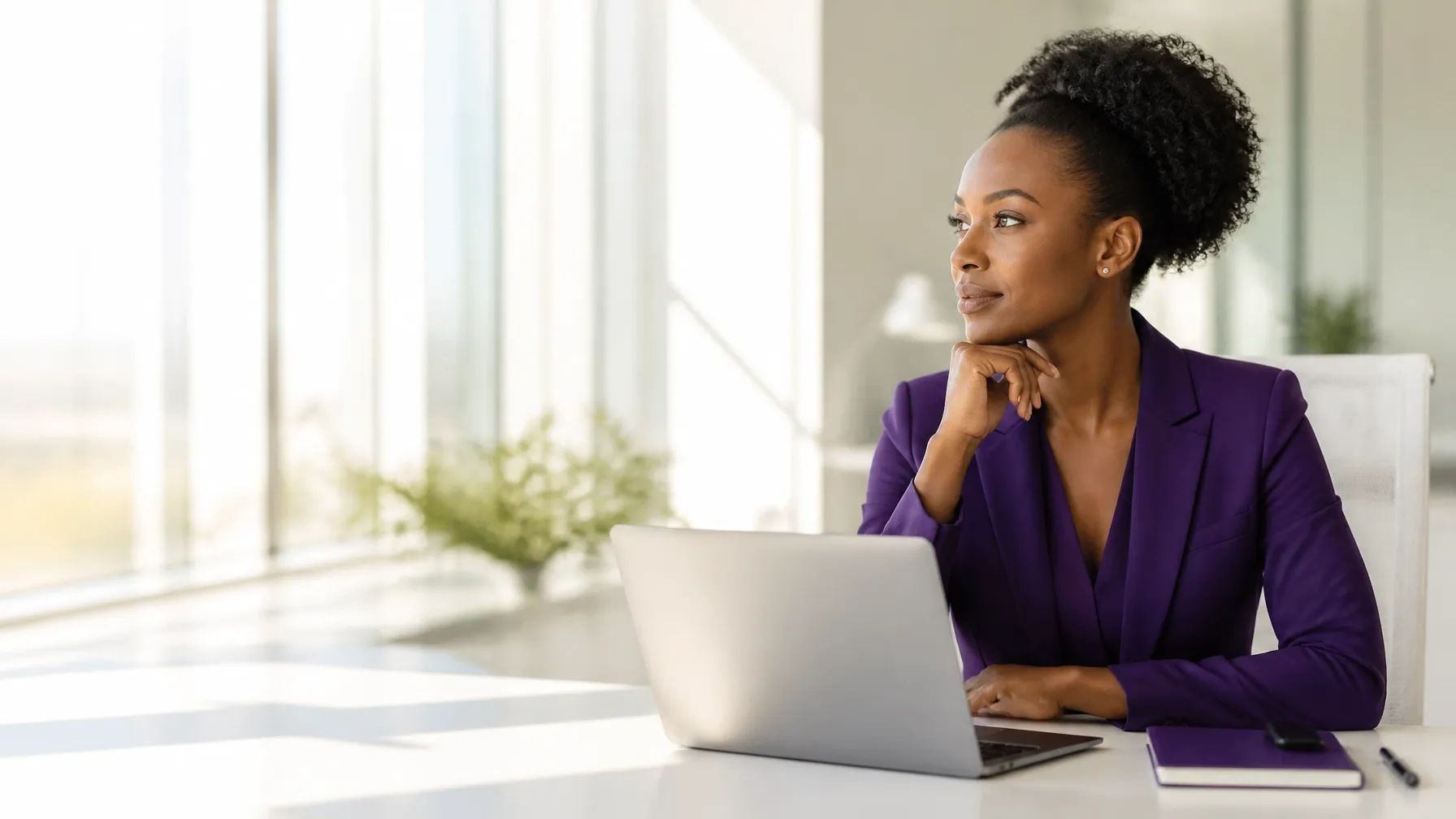 Professional Black woman in a purple blazer planning at a sunlit modern office desk with a laptop and notebook